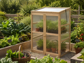 A small wooden greenhouse with clear plastic stands on a gravel path in a garden bed surrounded by leafy vegetables and tomato plants. Leading Landscapes Canterbury