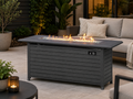 A dark gray outdoor fire pit storage table with a built-in firebox and visible flames, placed on a patio next to a light gray sofa with pillows. Leading Landscapes Canterbury