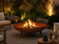A round metal fire pit bowl with a warm orange-brown finish, containing a bright fire, placed on a gravel patio in a landscaped garden setting with outdoor seating and warm ambient lighting. Leading Landscapes Canterbury