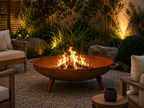 A round metal fire pit bowl with a warm orange-brown finish, containing a bright fire, placed on a gravel patio in a landscaped garden setting with outdoor seating and warm ambient lighting. Leading Landscapes Canterbury