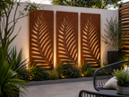 Three vertical rectangular metal wall panels with a leaf cutout pattern, mounted on a white wall in a landscaped outdoor patio setting with plants and warm uplighting. Leading Landscapes Canterbury
