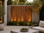 Modern outdoor patio scene with three tall decorative metal screen panels mounted on a concrete wall, backlit by warm ground lights. The scene includes landscaping, a small tree, a white outdoor sofa with a pillow, and a concrete coffee table in the foreground. Leading Landscapes Canterbury