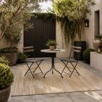 Outdoor patio with a round table and two chairs on a beige rug, surrounded by plants and a black gate. - Leading Landscapes Canterbury