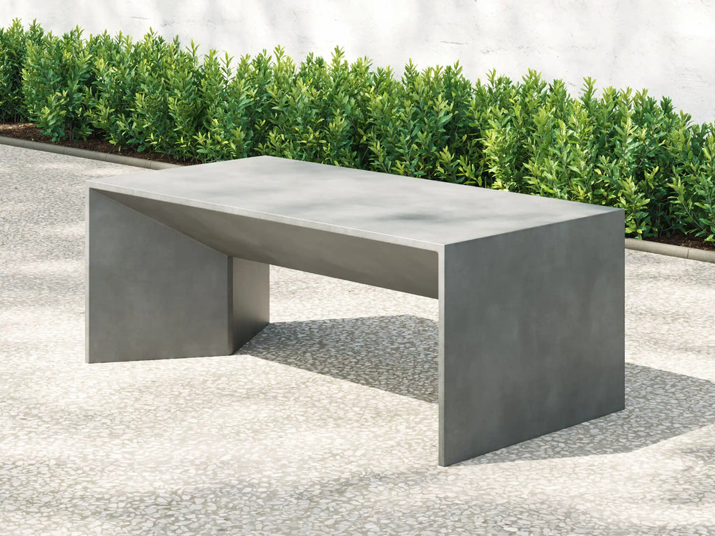 A modern rectangular outdoor bench with a matte gray concrete finish, positioned on a light gray paved surface with green hedges and a white wall in the background. Leading Landscapes Canterbury
