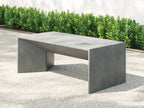 A modern rectangular outdoor bench with a matte gray concrete finish, positioned on a light gray paved surface with green hedges and a white wall in the background. Leading Landscapes Canterbury