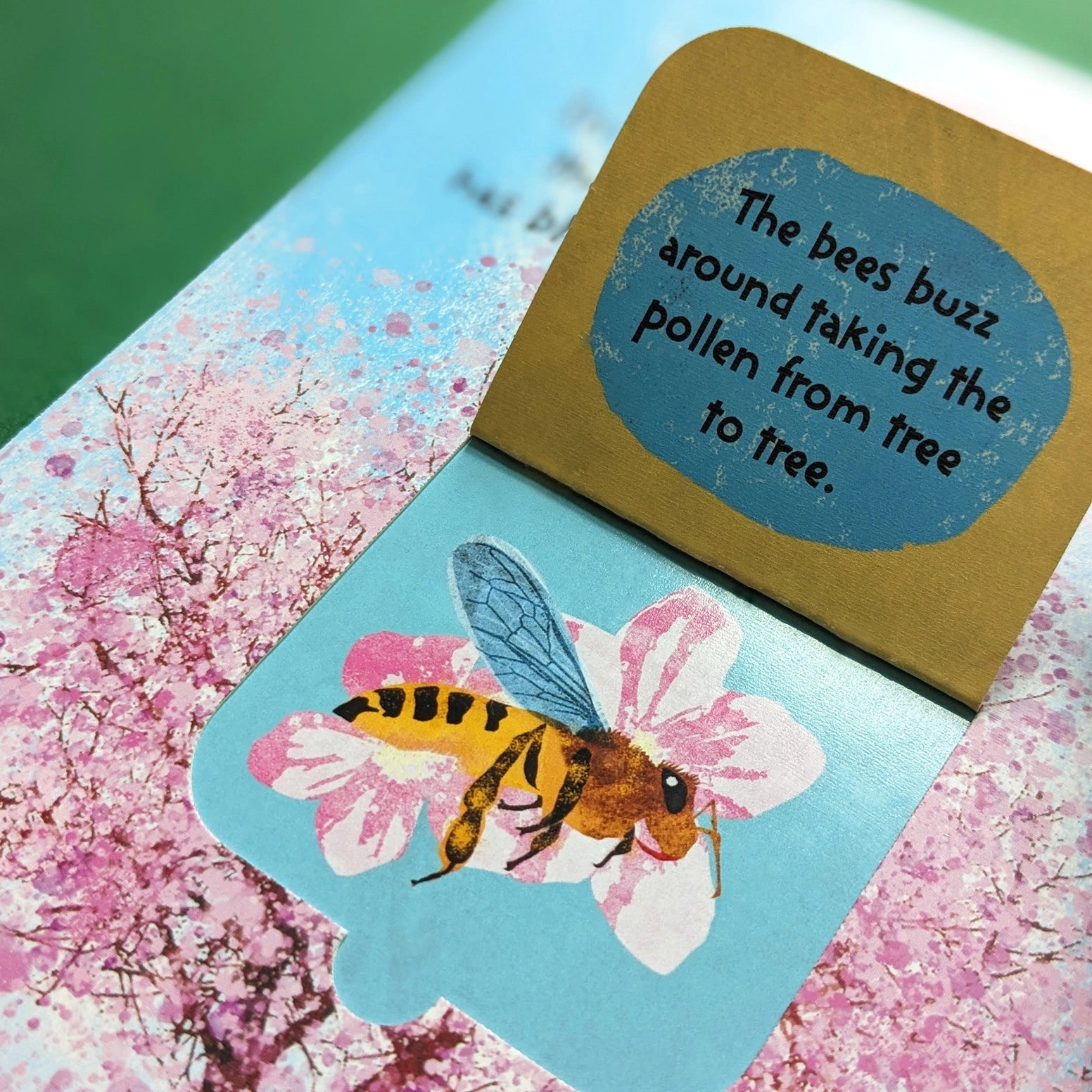 A close-up of a children's book or card featuring a bee on a pink flower, with a gold flap showing the text, 'The bees buzz around taking the pollen from the tree to tree.'.
