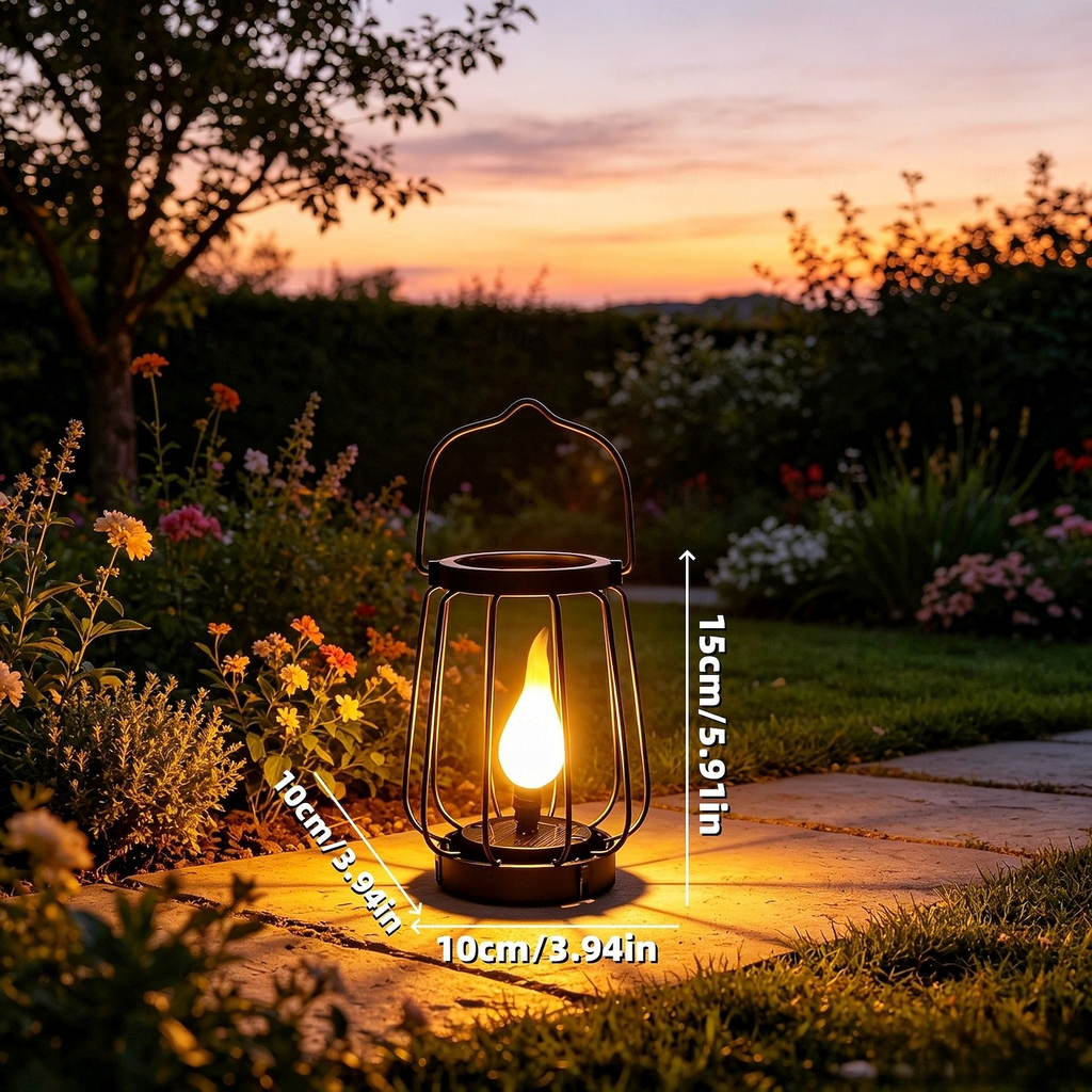 A black metal lantern-style outdoor light with a glowing yellow flame effect, placed on a stone path in a garden at sunset. White dimension labels show 15cm height and 10cm width.