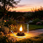 A black metal lantern-style outdoor light with a glowing yellow flame effect, placed on a stone path in a garden at sunset. White dimension labels show 15cm height and 10cm width.
