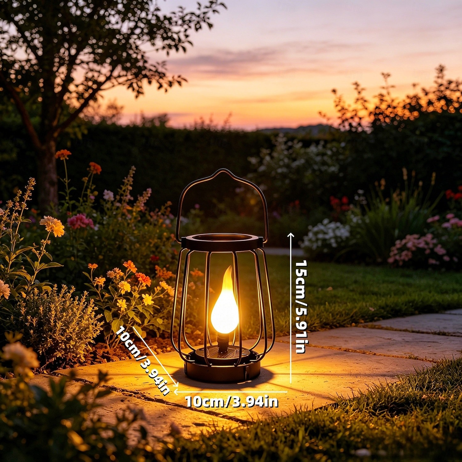 A black metal lantern-style outdoor light with a glowing yellow flame effect, placed on a stone path in a garden at sunset. White dimension labels show 15cm height and 10cm width.