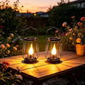 Two black metal wire lantern candles with glowing flame-shaped bulbs, placed on a wooden table in a garden at sunset. Solar lantern - Leading Landscapes Canterbury