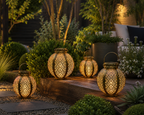 Four decorative metal lanterns with intricate cutout patterns, glowing with warm light, arranged on a wooden bench in a landscaped garden setting.