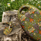 A decorative garden tool set featuring a large hand-painted green tin canister and two matching small hand tools with a William Morris-style floral pattern, displayed on a weathered tree stump with a blurred garden background.