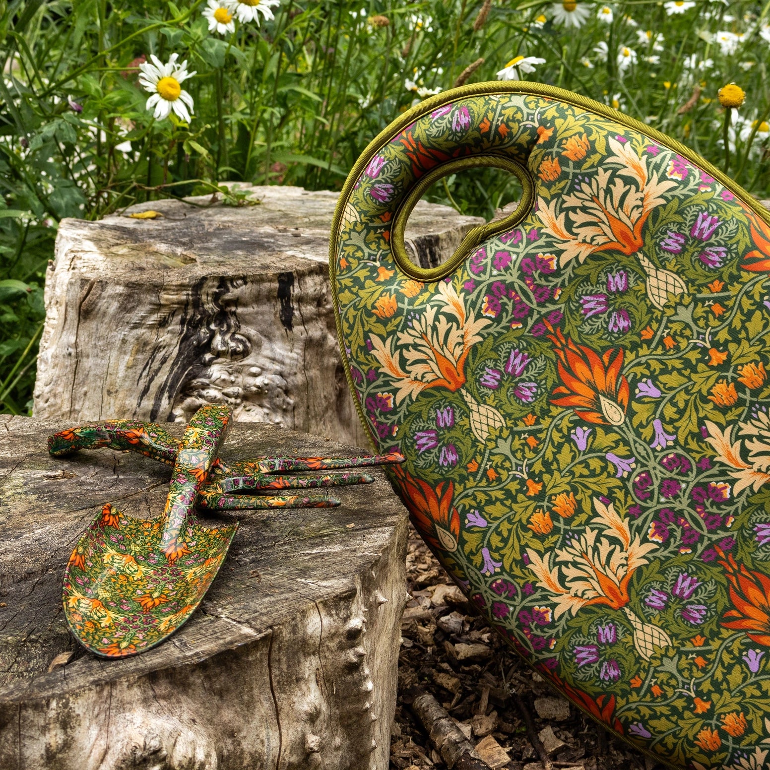 A decorative garden tool set featuring a large hand-painted green tin canister and two matching small hand tools with a William Morris-style floral pattern, displayed on a weathered tree stump with a blurred garden background.
